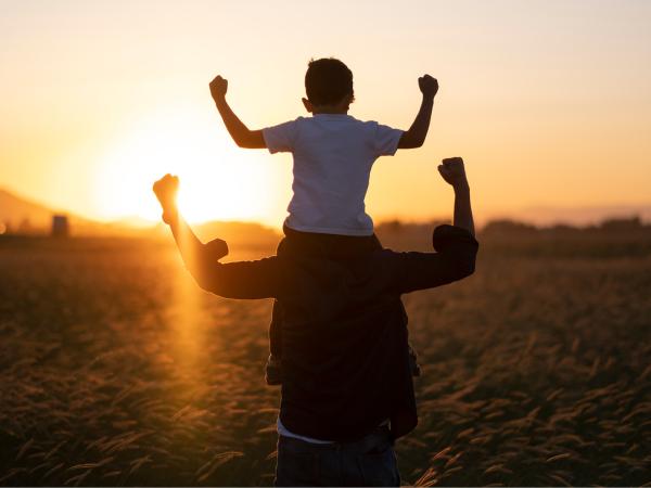 Child sat on an adult's shoulder walking towards the sun as it's setting in the distance