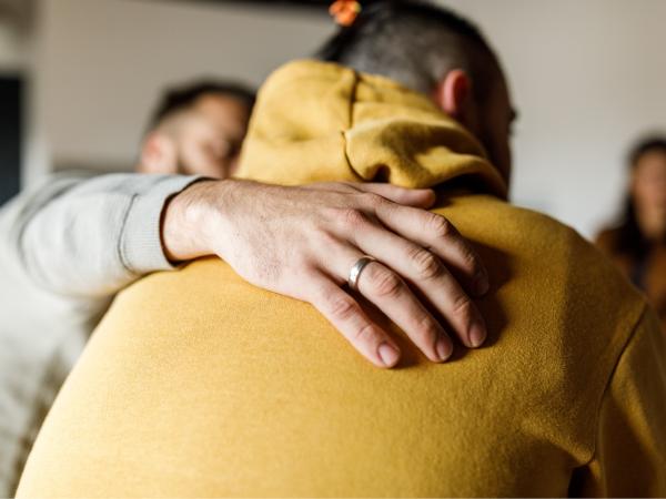 Two men with their backs to us, one has their arm around the other, comforting them