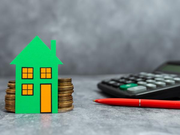 A green cardboard house resting on some stacked 2 pound coins on a table, plus a calculator and pen