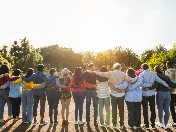 A group of 14 people standing in a row with their arms around each other, facing away from us 